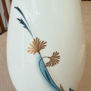 May include: A white ceramic vase featuring a smooth, rounded silhouette. The surface is decorated with a stylised design consisting of a curved, light blue brushstroke accent, dark blue leaf-like details, and two gold-coloured floral motifs.