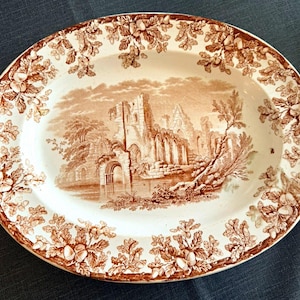 May include: An oval ceramic platter featuring a brown transferware pattern. The centre depicts a detailed landscape of stone ruins near a body of water. The wide rim is decorated with a repeating border of oak leaves and acorns in brown.