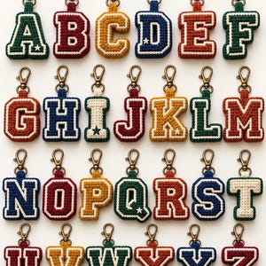 May include: A collection of twenty-six individual crochet letter keychains, each featuring a varsity-style block letter with a white border and a small star accent. Every keychain is attached to a bronze-toned metal lobster clasp hook.