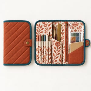 Peut inclure: Un étui de rangement en tissu matelassé orange brûlé avec une bordure bleu sarcelle et une fermeture à pression. L'intérieur présente une doublure à motif floral, des boucles élastiques pour stylos, un outil en bois et une poche zippée.