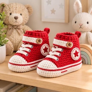 Könnte beinhalten: Ein Paar rot-weiße gehäkelte Babysneaker mit weißen Schnürsenkeln, einem weißen Riemen mit Holzknopf und einem runden Aufnäher an der Seite mit einem roten Stern. Die Schuhe haben eine strukturierte Strickoptik mit weißer Zehenkappe und Sohle.