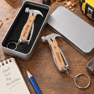 May include: Two multi-tool keychains featuring a miniature hammer head and light wood handle. One tool is engraved with "Best Dad" and rests in a silver metal tin, while the other is engraved with "James" and lies on a wooden surface.