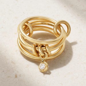 May include: A gold ring featuring multiple interlocking smooth bands of varying thicknesses. A small, circular charm with a single clear, round-cut gemstone hangs from the center of the bands. The interior of the ring is stamped with the text SK 18K.