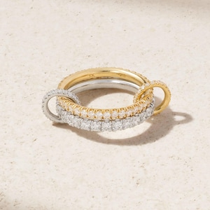 Pode incluir: Um conjunto de anéis entrelaçados composto por uma banda de ouro lisa com as letras SK, uma banda de prata e dois anéis tipo eternity cravejados com pequenas pedras claras. A peça combina tons dourados e prateados com brilhos.