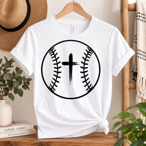 Puede incluir: Una camiseta blanca de manga corta con un gran gráfico negro de una pelota de béisbol que tiene una cruz de estilo pincelada en el centro. La camiseta tiene cuello redondo y se muestra colgada en una percha de madera.