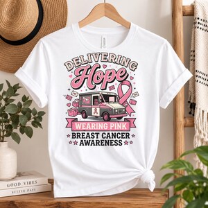 May include: A white short-sleeve t-shirt featuring a graphic of a mail truck with a pink ribbon, surrounded by envelopes and hearts. The text reads: Delivering Hope, Wearing Pink, Breast Cancer Awareness. The design uses pink, grey, and black colours.