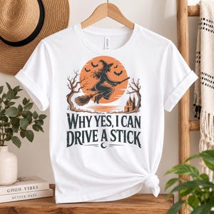 May include: A white t-shirt featuring a graphic of a witch flying on a broomstick in front of a large orange moon. The design includes silhouettes of bats, bare trees, and a castle. Below the graphic, the text reads: WHY YES, I CAN DRIVE A STICK.