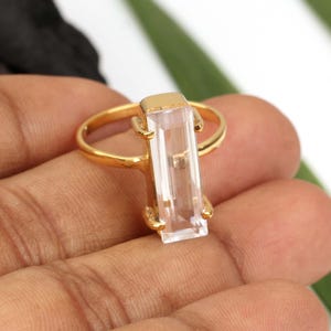 Könnte beinhalten: Ein goldfarbener Ring mit einem einzelnen, länglichen, rechteckig geschliffenen, klaren Edelstein, der vertikal gefasst ist. Der Stein wird von vier kleinen Krappen auf einer dünnen, polierten goldfarbenen Metallschiene gehalten.
