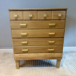 May include: A light brown wooden dresser featuring four large drawers with horizontal gold metal pulls and a top row of four smaller drawers accented with round, textured gold knobs. The piece stands on four tapered wooden legs.