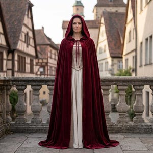 May include: A floor-length, deep red velvet hooded cloak with a tie closure at the neck, worn over a long, cream-coloured sleeveless dress. The cloak features a full hood and drapes down to the floor, covering the dress underneath completely.