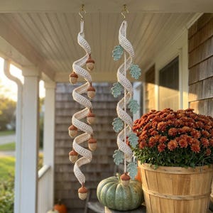 May include: Two hanging spiral decorations made of cream-colored yarn. One spiral features small crocheted acorns in brown and tan, while the other spiral is adorned with small crocheted oak leaves in a soft sage green color. Both hang from gold hooks.