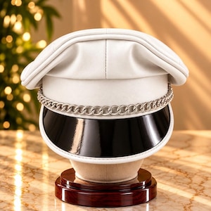 Pode incluir: Um boné de estilo militar branco com um acabamento liso e mate. O chapéu inclui uma pala preta brilhante proeminente e uma tira decorativa de corrente de metal prateado que repousa horizontalmente sobre a base frontal da coroa.