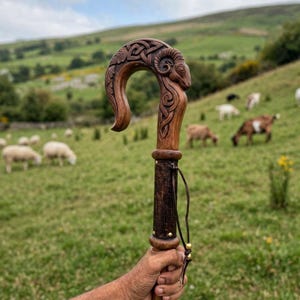 May include: A wooden shepherd's crook featuring a hand-carved ram's head at the top, detailed with intricate Celtic knotwork patterns. The shaft is made of dark wood, accented with small brass studs and a thin leather cord tied around the handle.