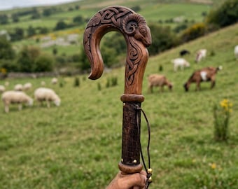 May include: A wooden shepherd's crook featuring a hand-carved ram's head at the top, detailed with intricate Celtic knotwork patterns. The shaft is made of dark wood, accented with small brass studs and a thin leather cord tied around the handle.