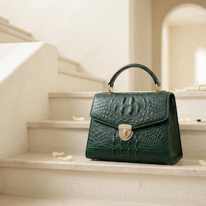 Peut inclure: Un sac à main vert foncé avec un motif texturé en peau de crocodile, doté d'une poignée supérieure structurée, d'un rabat et d'une fermeture à pression en métal doré. Le sac présente une forme trapézoïdale et des finitions en métal doré.