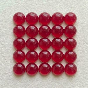 Könnte beinhalten: Eine Sammlung von fünfundzwanzig kreisförmigen, durchscheinenden roten Cabochons, die in einem fünf-mal-fünf-Raster angeordnet sind. Jedes einzelne Stück hat eine glatte, polierte, gewölbte Oberfläche, die Licht reflektiert.