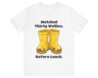 May include: A white short-sleeve t-shirt featuring a graphic of two yellow rubber boots covered in brown mud at the base. The text above the boots reads "Matched Thirty Wellies." and the text below the boots reads "Before Lunch."