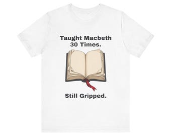 May include: A white short-sleeve t-shirt featuring a graphic of an open book with a red ribbon bookmark. The text above the book reads Taught Macbeth 30 Times. and the text below the book reads Still Gripped. in a simple black font.