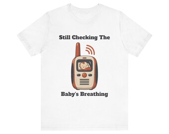 May include: A white short-sleeve t-shirt featuring a graphic of a baby monitor with a sleeping infant on the screen and red signal waves. The text above the monitor reads Still Checking The and below the monitor reads Baby's Breathing.