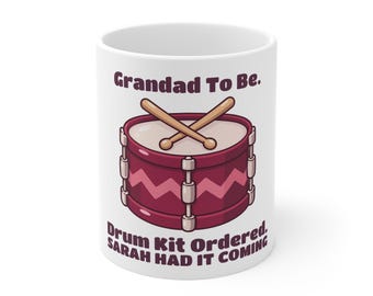May include: A white ceramic mug featuring a maroon drum with two crossed wooden drumsticks. Above the drum is the text "Grandad To Be." and below the drum is the text "Drum Kit Ordered. SARAH HAD IT COMING" in dark maroon lettering.