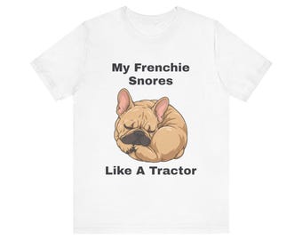 May include: A white short-sleeve t-shirt featuring a graphic of a sleeping tan French Bulldog curled into a ball. Above the dog, the text reads My Frenchie Snores, and below the dog, the text reads Like A Tractor in dark gray lettering.
