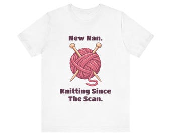 May include: A white short-sleeve t-shirt featuring a graphic of a pink ball of yarn with two crossed wooden knitting needles. The text above reads New Nan. and the text below reads Knitting Since The Scan. in dark brown lettering.