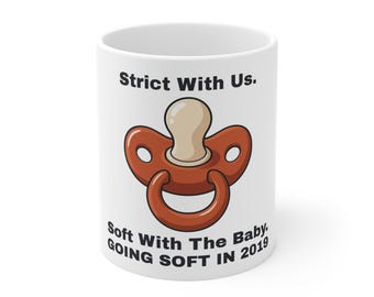 May include: A white ceramic mug featuring a graphic of a brown baby pacifier in the center. The text above the pacifier reads Strict With Us. The text below the pacifier reads Soft With The Baby. GOING SOFT IN 2019 in black lettering.
