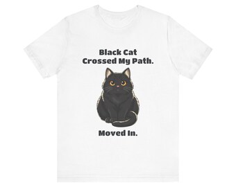 May include: A white short-sleeve t-shirt featuring a graphic of a black cat with yellow eyes. The text above the cat reads Black Cat Crossed My Path. and the text below the cat reads Moved In. The design is centered on the front of the shirt.