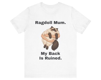 May include: A white short-sleeve t-shirt featuring a graphic of a fluffy Ragdoll cat being held. Above the cat, the text reads "Ragdoll Mum." and below the cat, the text reads "My Back Is Ruined." The cat has cream fur with dark brown points.