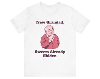 May include: A white t-shirt featuring a graphic of an older person wearing a pink cardigan, winking and holding a finger to their lips. The text above reads New Grandad. and the text below reads Sweets Already Hidden. in dark brown.