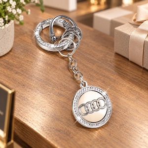 May include: A silver-toned metal keychain featuring a circular charm with the Audi four-ring logo in the center. The charm and the main ring are both bordered by small, clear, sparkling crystals. The keychain includes a chain and a spring-loaded clasp.