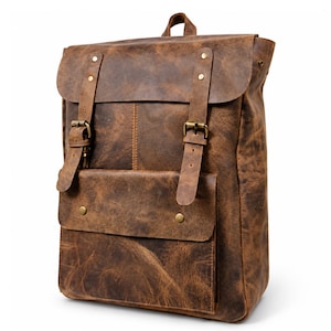 Puede incluir: Una mochila de cuero marrón con una solapa superior plegable y dos correas verticales aseguradas con hebillas de metal en tono latón. El frente incluye un bolsillo rectangular con dos botones a presión de latón y un asa superior.