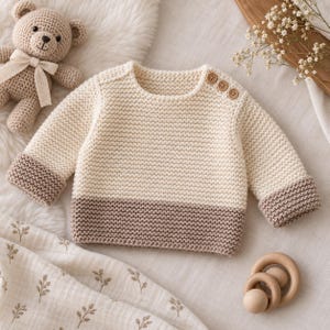 Könnte beinhalten: Ein langärmeliger Strickpullover mit einem cremefarbenen Oberteil und einem braunen Unterteil. Das Kleidungsstück hat einen Rundhalsausschnitt mit drei kleinen Holzknöpfen an der linken Schulter und passende braune Bündchen an den Ärmeln.