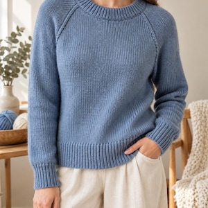 Könnte beinhalten: Ein langärmeliger Strickpullover in Staubblau mit geripptem Rundhalsausschnitt, gerippten Bündchen und geripptem Saum. Der Pullover hat Raglanärmel und ein strukturiertes Strickmuster, kombiniert mit einer hellbeigen weiten Hose.