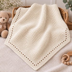 May include: A cream-colored knitted blanket featuring a textured garter stitch pattern and a decorative border with evenly spaced eyelet holes. The square-shaped textile is draped over a woven basket, showcasing its soft, uniform knit construction.