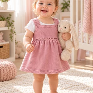 May include: A toddler wearing a dusty rose knitted sleeveless dress over a plain white short-sleeved shirt. The dress features a square neckline, a decorative eyelet row at the waist, and a flared skirt. The toddler holds a plush cream-colored rabbit.