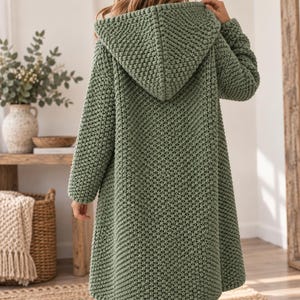 Puede incluir: Un cárdigan con capucha de color verde salvia y mangas largas, con un patrón de tejido grueso y texturizado. La prenda llega hasta la mitad del muslo e incluye una capucha grande integrada. El tejido es uniforme en la espalda y mangas.