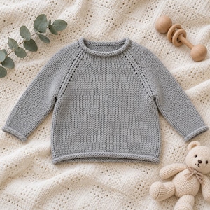 Puede incluir: Un suéter de punto gris claro de manga larga con cuello redondo y mangas raglán. La prenda está confeccionada con un patrón de punto jersey visible, acabado con bordes enrollados en el cuello, los puños y el dobladillo para un aspecto sencillo.