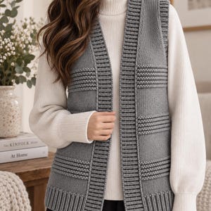 Puede incluir: Un chaleco de punto gris sin mangas con una tapeta frontal acanalada vertical y bandas horizontales texturizadas en el cuerpo. El chaleco se lleva sobre un suéter blanco de manga larga con cuello alto, creando un estilo de capas.