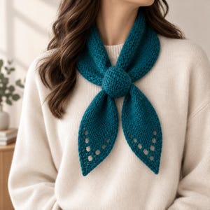 Op de afbeelding: Een blauwgroene gebreide halsdoek met een centraal rond knoopdetail en twee puntige uiteinden met een decoratief opengewerkt kantpatroon langs de randen. Het accessoire wordt gedragen over een crèmekleurige gebreide trui met lange mouwen.