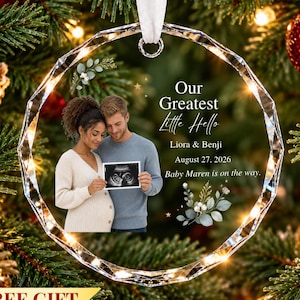 May include: A circular, faceted glass Christmas ornament featuring a photo of a couple holding an ultrasound image. Text reads: Our Greatest Little Hello, Liora & Benji, August 27, 2026, Baby Maren is on the way. Includes a white ribbon.