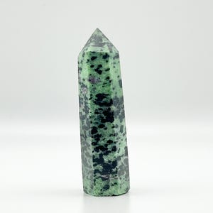 May include: A green and black zoisite crystal point stands upright against a white background. The polished crystal has a faceted shape and a pointed top. The stone's surface is speckled with dark green and black spots.