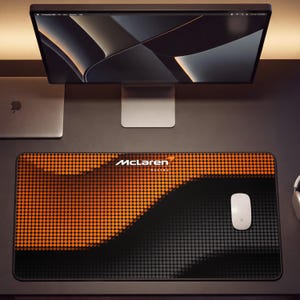 May include: A rectangular desk mat featuring a gradient pattern of orange and black dots. The McLaren Racing logo is printed in white text at the top center. A white computer mouse rests on the right side of the mat, which is placed on a desk.