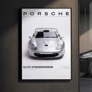 May include: A framed wall poster featuring a silver Porsche 911 GT1 Strassenversion centered on a white background. The text PORSCHE appears at the top, with 911 GT1 STRASSENVERSION and technical specifications printed at the bottom of the print.