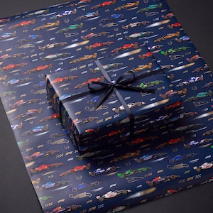 May include: A rectangular gift box wrapped in dark blue paper featuring a repeating pattern of Formula 1 race cars, checkered flags, and the F1 logo. The box is tied with a black satin ribbon tied in a bow on top, resting on a matching sheet of paper.