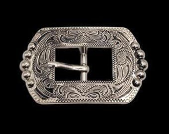 May include: Silver-toned belt buckle with an ornate engraved design and a rectangular shape. The buckle has a smooth, polished finish and features a row of small beads along the edge.