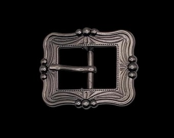 May include: A silver-toned metal belt buckle with an ornate design. The buckle has a rectangular shape with rounded corners and a detailed floral pattern.