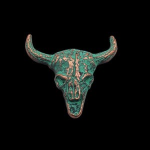 May include: A green and copper colored bull skull charm. The skull is detailed with a textured surface and has a distressed look.