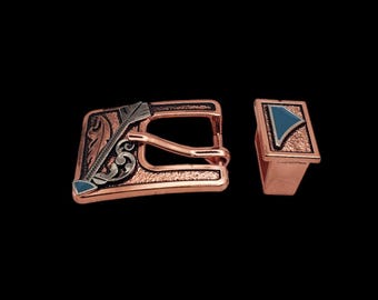 May include: A copper-colored belt buckle with an intricate design featuring a silver arrow and floral patterns. The buckle has a blue stone inlay and comes with a matching keeper.