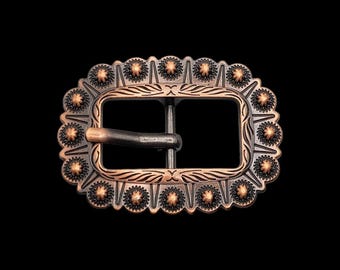 Könnte beinhalten: Eine ovale Gürtelschnalle mit kupferfarbenem Finish. Der Rahmen zeigt ein sich wiederholendes Muster aus kleinen, erhabenen sternartigen Nieten am Außenrand sowie gravierte blattartige Details, die die zentrale rechteckige Öffnung und den Dorn umgeben.